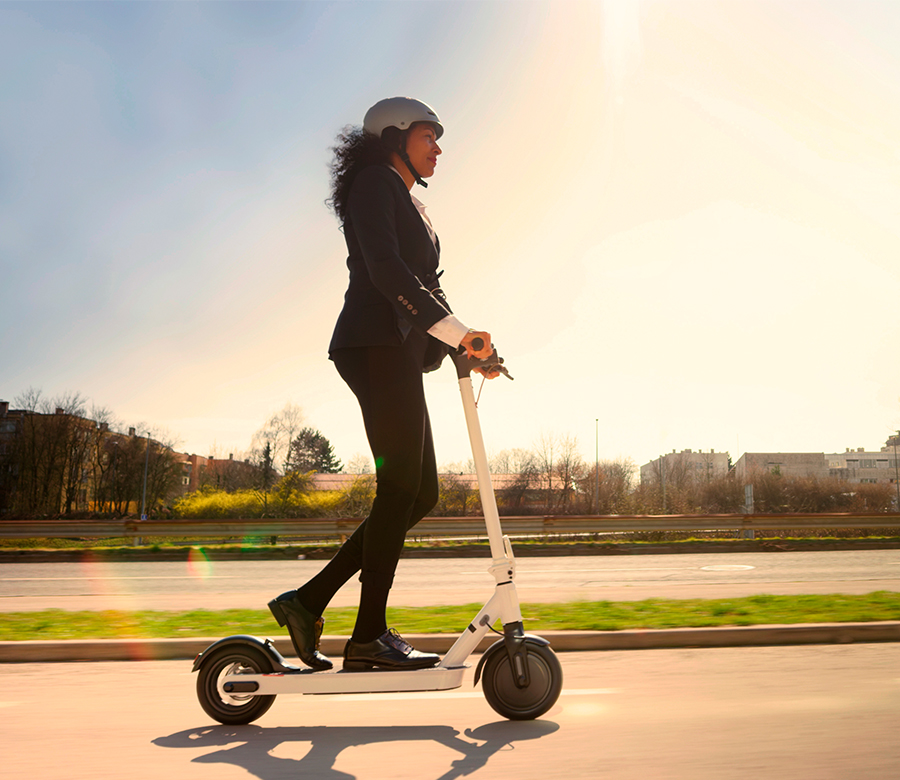 Imatge de perfil d'una dona en un patinet elèctric blanc, de camí a l'oficina. Vesteix un vestit fosc de negocis i un casc de protecció blanc. S'apropa a la seu de Gabinet Senia & Associats a Lloret de Mar, una empresa especialitzada en assegurances de patinet MGS i gestió de finques